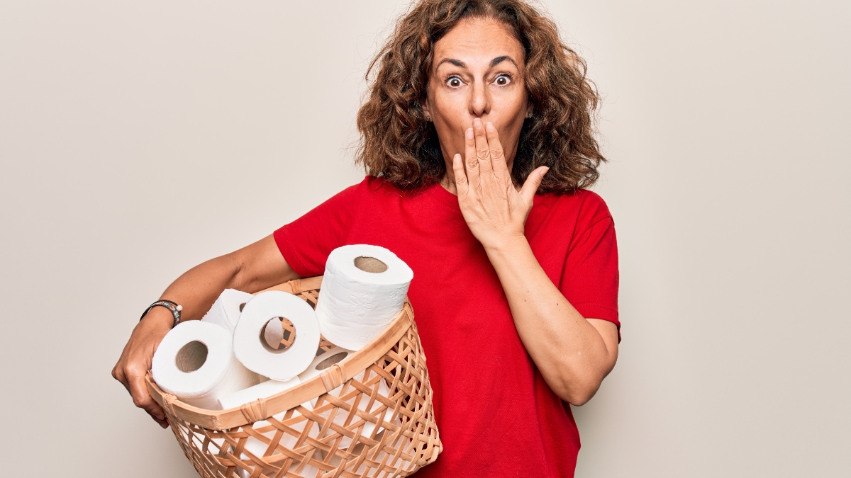 Frau mittleren Alters hält einen Weidenkorb mit vielen Toilettenpapierrollen vor weißem Hintergrund und bedeckt den Mund mit der Hand.