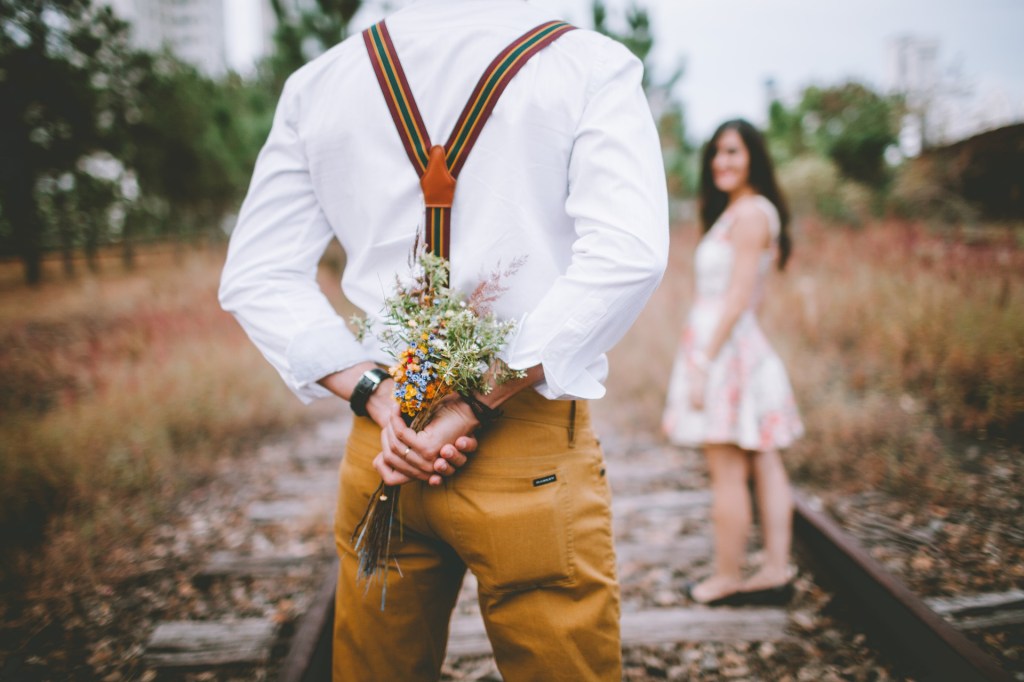 Ein Paar steht auf Bahnschienen und der Mann hält Blumen hinter dem Rücken versteckt.