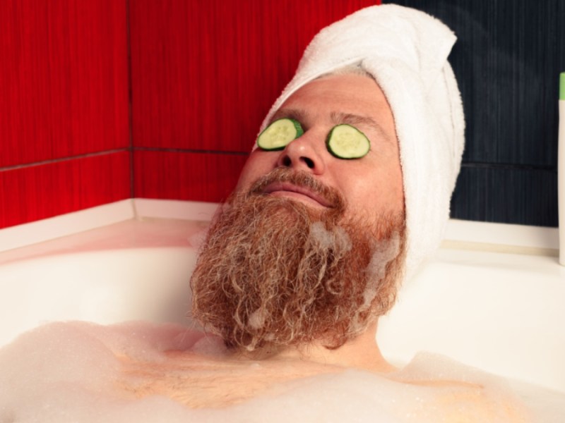 Niedlicher bärtiger Mann, der ein Bad nimmt, den Kopf in ein Handtuch gewickelt und Gurkenscheiben auf den Augen.