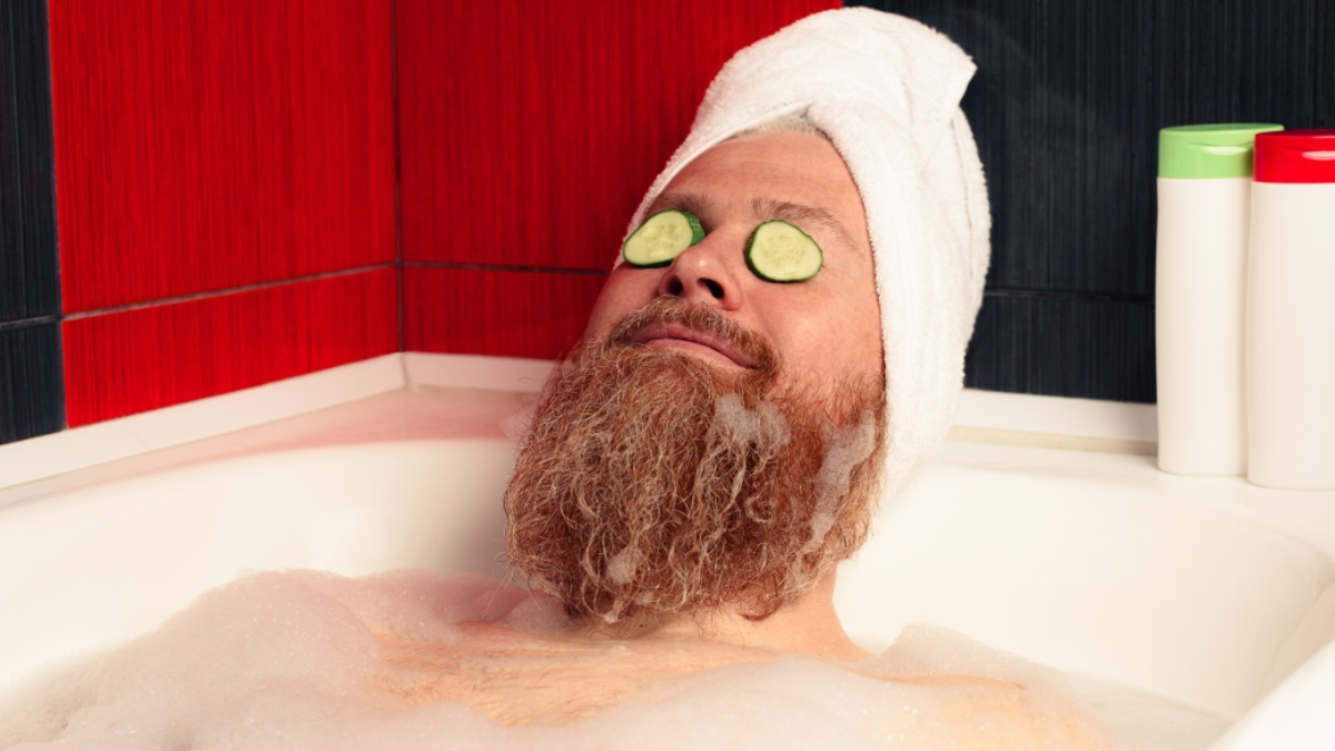 Niedlicher bärtiger Mann, der ein Bad nimmt, den Kopf in ein Handtuch gewickelt und Gurkenscheiben auf den Augen.