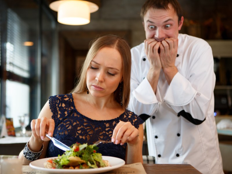 Ein Koch kaut sich wütend die Nägel, während er einem Gast beim Essen zusieht.