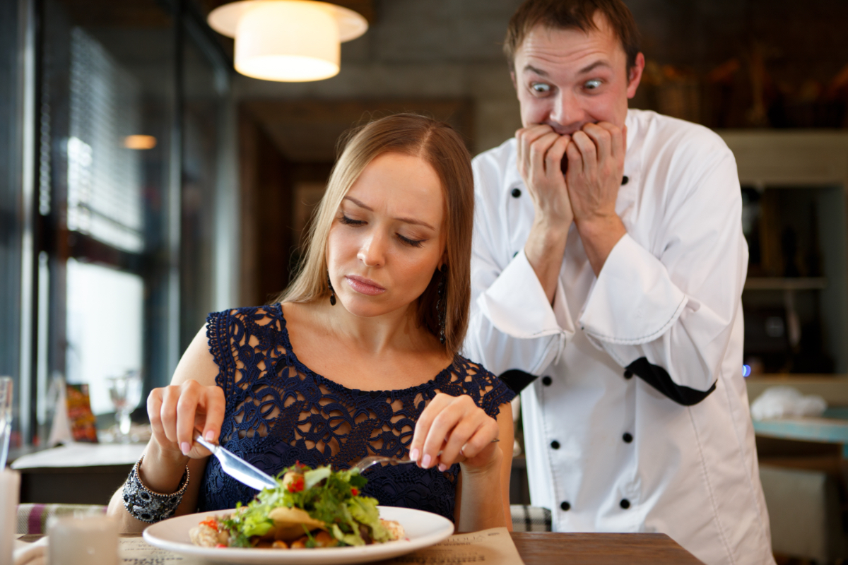Ein Koch kaut sich wütend die Nägel, während er einem Gast beim Essen zusieht.