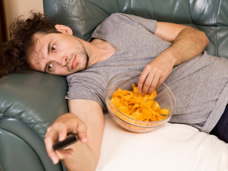Ein fauler Mann liegt auf der Couch, schaut Fernsehen und isst Chips.