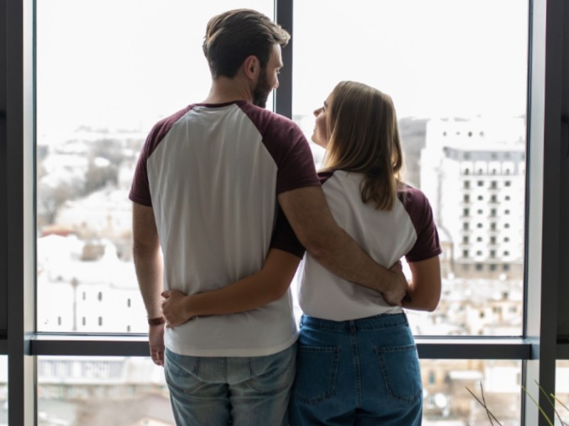 Junges Paar, das sich umarmt, steht am Fenster und genießt die Aussicht von seiner neuen Wohnung.