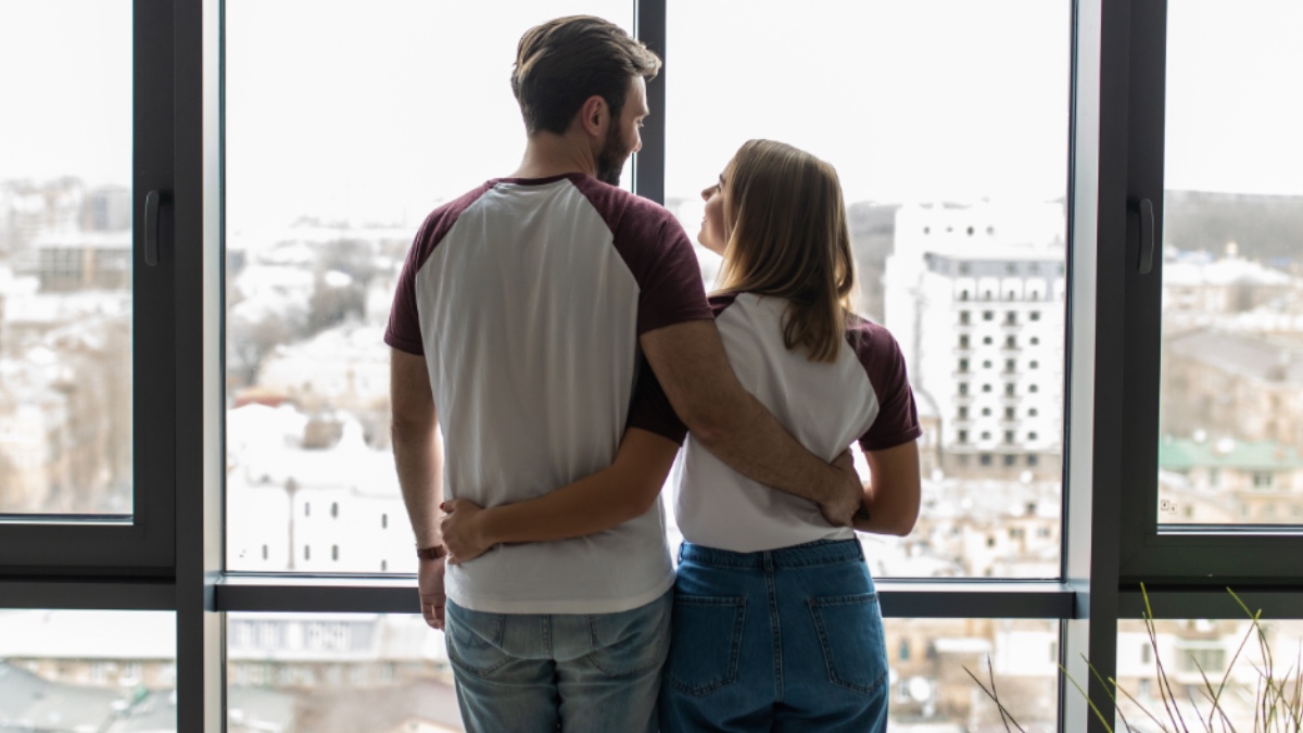 Junges Paar, das sich umarmt, steht am Fenster und genießt die Aussicht von seiner neuen Wohnung.