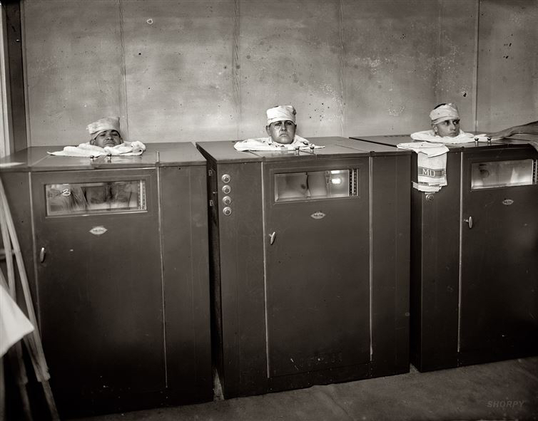 Drei Männer stecken in Physiotherapie-Boxen, ca. 1920.