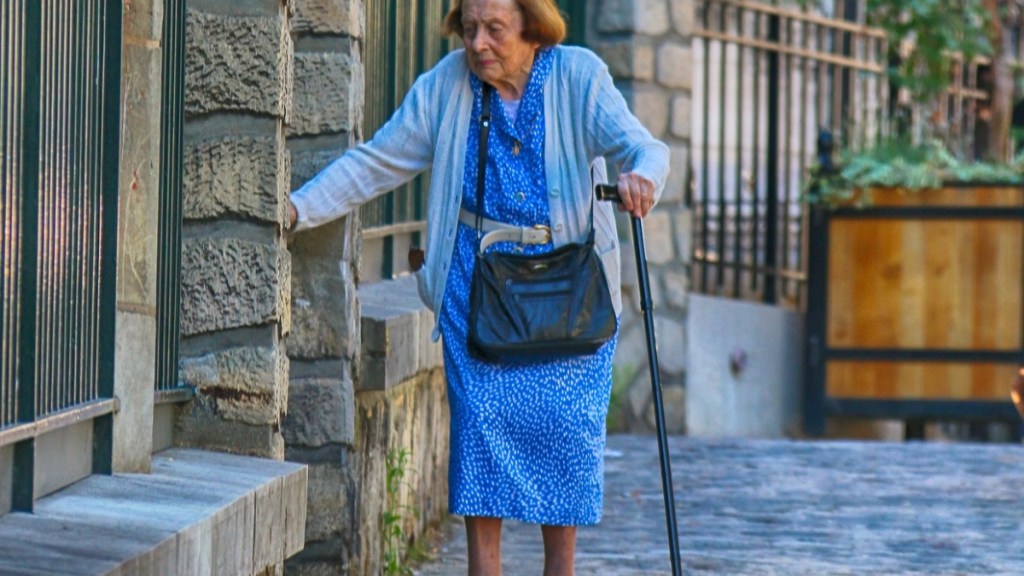 Eine alte Frau mit Gehstock stützt sich an einer Mauer ab.